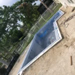 Under-construction in-ground swimming pool with a blue liner, white fence, and surrounding sandy ground in a residential yard with palm trees.