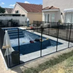 Under-construction in-ground pool with blue tile interior surrounded by black mesh safety fence; worker in the background and construction materials nearby.