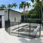 Under-construction rectangular in-ground pool in a sunny tropical backyard, enclosed by a black mesh fence; palm trees and a white building with a slanted awning nearby.