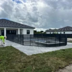 Construction worker in a neon shirt installs a black mesh safety fence around a rectangular inground pool in a modern white house backyard under a cloudy sky.