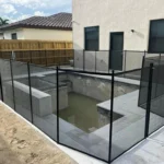 Rectangular backyard pool under construction, surrounded by black mesh safety fence and posts, with a beige house and sliding door in the background.
