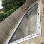 Rectangular pool under construction with a black safety net draped over white coping in a fenced backyard.