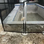 Backyard in-progress: black metal and glass pool fence surrounds an empty in-ground pool with construction debris on the dirt ground outside the fence.