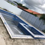 Under-construction in-ground swimming pool with blue tile edge and safety net, in a fenced backyard neighborhood.