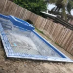 Rectangular in-ground pool under construction, covered with a blue safety net, in a backyard with a wooden fence and palm tree in the background.
