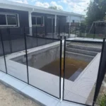 Black metal-framed mesh fence surrounding an empty in-ground pool pit in a backyard patio area. The house and outdoor toys are visible in the background.