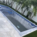 Long rectangular pool under construction, covered with blue safety netting; white tile surround and palm trees along a white fence in the yard.