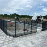 Construction site of an in-ground pool surrounded by a black safety fence; worker in orange shirt stands inside the pool.