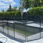 Rectangular in-ground pool under construction, enclosed by a tall black mesh safety fence in a modern backyard with hedges and trees in the background.