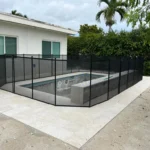 Backyard with an empty rectangular in-ground pool surrounded by a black mesh safety fence on a light tiled patio.