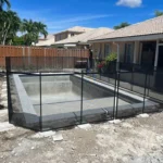 Vacant in-ground pool under construction surrounded by a black safety fence in a sunny backyard.