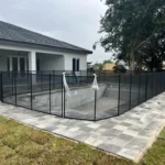 In-ground pool under construction surrounded by a black metal-frame glass panel safety fence adjacent to a modern white house with a covered patio and gray paver patio stones.