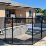 Black metal-framed mesh pool fence surrounding an empty in-ground pool in a backyard, with a beige house and blue sky in the background.