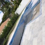 Blue protective netting covering a rectangular sunken pool area on a marble patio, with palm trees and a hedge in the background