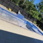 In-ground pool under construction with blue safety netting, blue-tiled steps, and a raised spa area, bordered by a beige paver patio and a tall black fence.