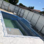 Under-construction in-ground pool with a blue safety net covering the water, surrounded by a white privacy fence and tiled deck.
