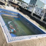 Under-construction rectangular in-ground pool with blue tile border and blue safety netting in a sandy yard near houses.
