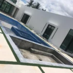 Modern white residence with large glass doors surrounding an empty blue-tiled pool under construction; palm trees in the background.