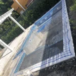 Blue safety mesh fencing surrounds a concrete pool frame under construction, with a gravel area and green hedge nearby.